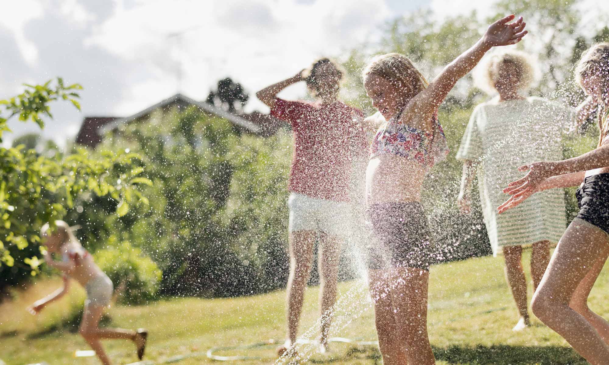 Barn och vuxna leker ute i en vattenspridare en solig sommardag